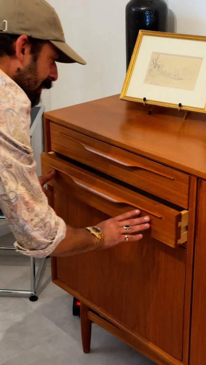 Malek Youssef inspectant une commode en bois teck, détail sur les tiroirs et un dessin encadré.