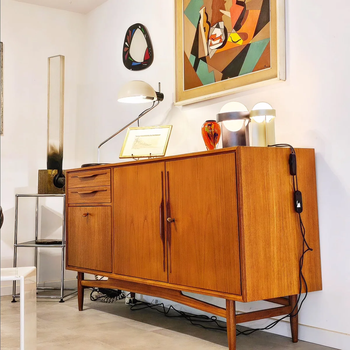 Buffet vintage en teck avec des luminaires, un tableau abstrait et des objets décoratifs sur fond blanc.
