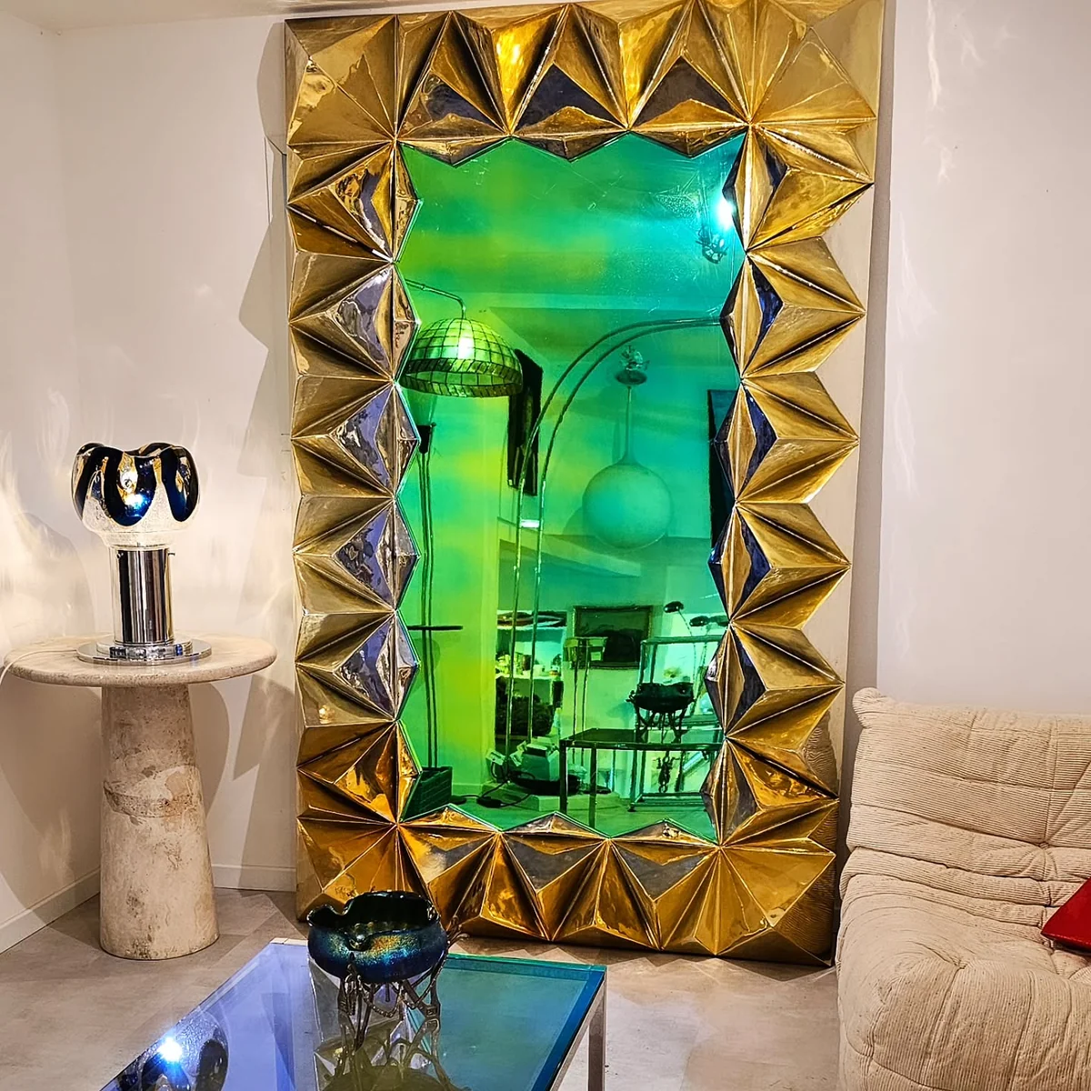 Miroir monumental doré à l'or et à facettes, reflétant la lumière verte et des meubles.