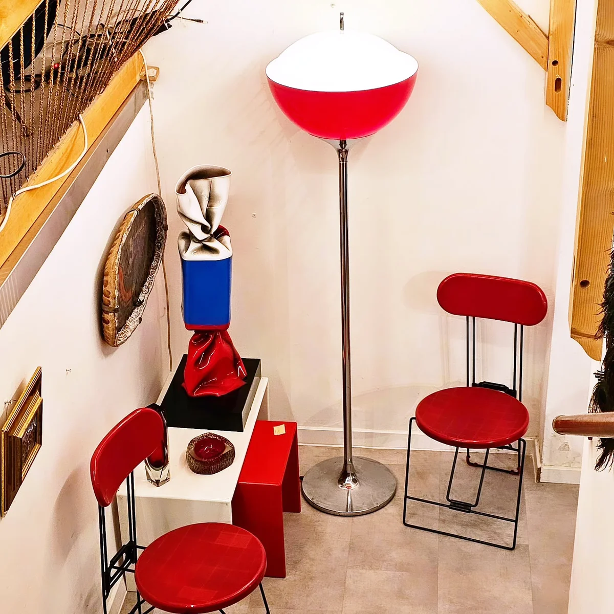 Un lampadaire rouge et blanc volumineuse à côté d'une sculpture originale et de chaises rouges.