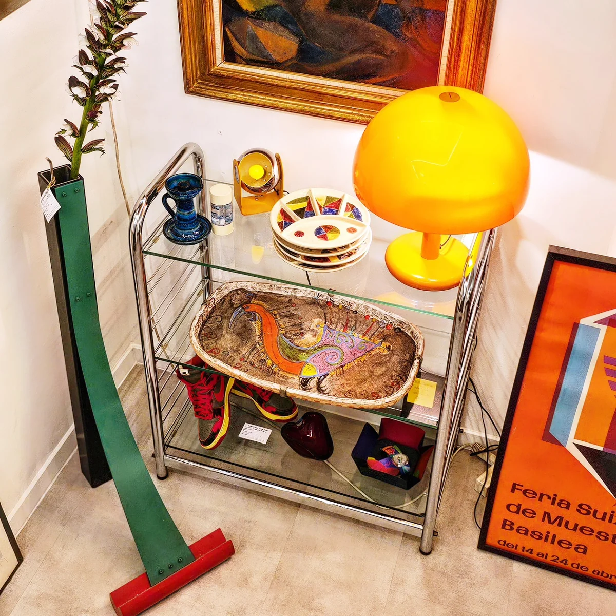 Détail d'une étagère en verre avec une lampe jaune, un plat décoratif et une affiche.