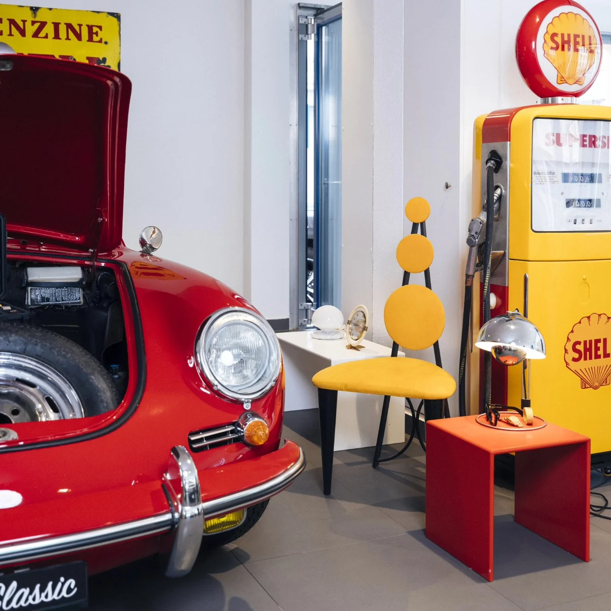 Voiture rouge rétro devant une pompe à essence Shell jaune, avec une chaise et une lampe design.